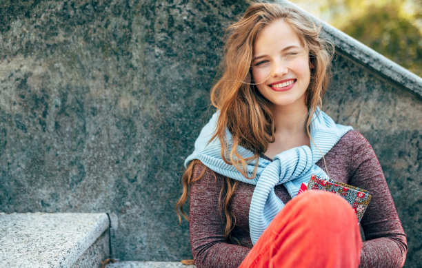 一名白人女性户外肖像坐在街上的楼梯上休息。快乐的年轻女学生带着健康的微笑看着镜头，坐在大学校园的旁边。图片下载