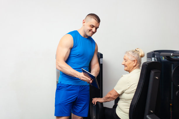 一个年轻英俊的教练向年长的女人展示如何进行锻炼，在平板上的图片。当前的体育技术。教练记录结果。复制空间，白色背景。微笑图片下载