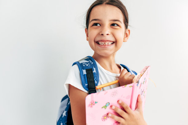 一个带着蓝色背包的快乐微笑女学生的特写照片在她的笔记本上写着什么，在浅灰色的工作室背景上摆姿势时看着一边。图片下载