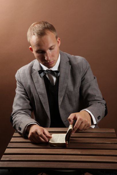 一个英俊，自信的商人，穿着西装，独自坐在棕色背景上读着一本书。在一张木桌旁边图片下载