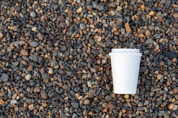 一个白色的纸杯咖啡或茶躺在岩石岸边，卵石图片下载