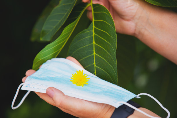 一张裁剪过的照片，一名女子手持一个医用口罩，口罩上有黄色的花朵，背景是一片绿叶。图片下载