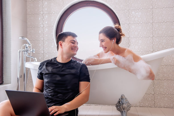 年轻男子坐在现代浴缸旁，把笔记本电脑放在膝盖上，和美女一起听音乐。男孩把耳机递给了泡在白色泡沫浴缸里的女孩。相爱的夫妻一起欢笑图片下载
