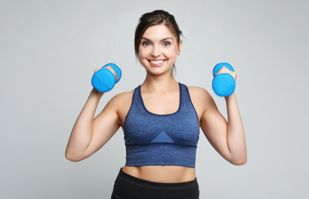 一张健康健康的年轻女子微笑着把哑铃举在肩上的照片。运动与人的概念图片下载
