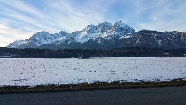 在一个阳光明媚的冬日夜晚，一辆电动汽车的驾驶窗向外移动，前面是白雪覆盖的阿尔卑斯山脉。柏油山路上的第一人称视角。图片下载