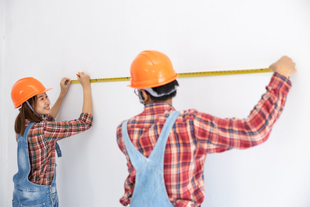 年轻的亚洲夫妇穿着格子衬衫和熊布和橙色的帽子工作和帮助测量卷尺和在白墙上标记铅笔团队夫妇室内装修的概念图片下载