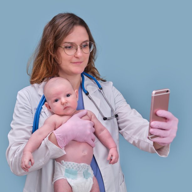 儿科医生拿着手机和新生儿，蓝色工作室背景。穿着制服的快乐护士带着孩子和手机图片下载
