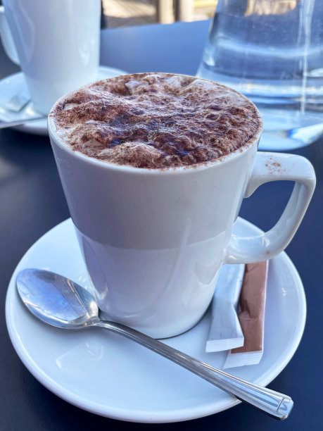 餐厅场景的图片，桌上有白色的杯子和托盘，上面撒着泡沫的热巧克力，上面撒着可可粉，巧克力粉涂在牛奶上，托盘里有茶匙和糖袋，聚焦前景图片下载