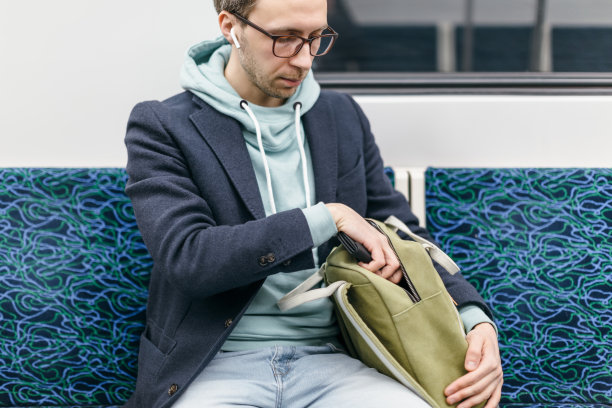 裁剪过的照片上，一个穿着蓝色外套、戴着眼镜、耳朵里戴着无线耳机的嬉皮士坐在那里，从绿色背包里掏出钱包。图片下载