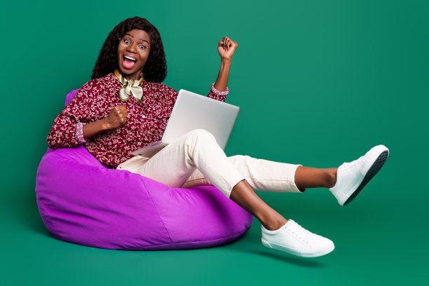 照片光泽幸运波浪深色皮肤女士穿着印花衬衫坐豆袋手持小工具上升拳头孤立的绿色背景图片下载