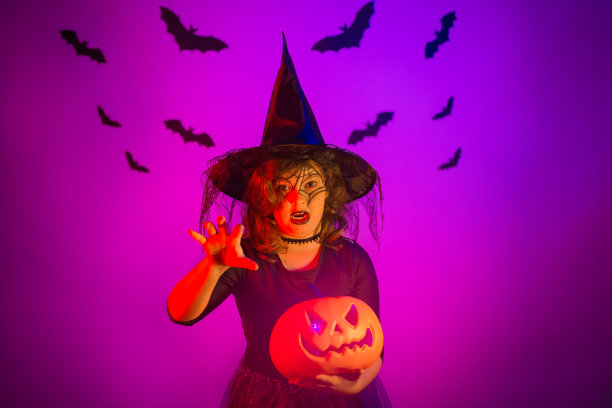 一个十几岁的女孩穿着万圣节女巫服装，脸上画着南瓜和蜘蛛网。工作室在美丽的霓虹灯紫色照明。万圣节快乐概念图片下载