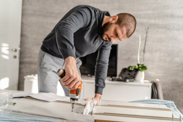 一个有胡子的男人独自在家里组装自组装家具，拿着电动螺丝刀螺丝和拼装件-半长正面DIY概念真人复制空间图片下载