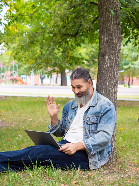 一名中年男子在线视频通话时的垂直照片。他坐在城市公园的草地上，手里拿着笔记本电脑。他挥着手——你好图片下载