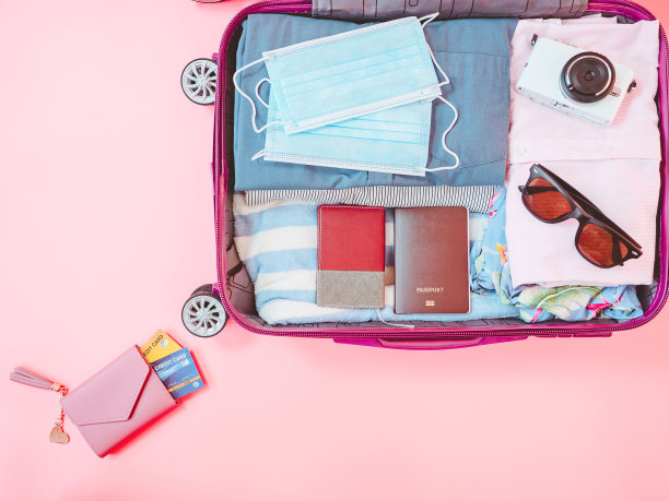 粉红色背景上有衣服和配件的手提箱包。伴随冠状病毒大流行的暑假旅行。图片下载