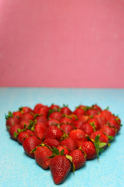 一堆新鲜的红草莓，绿色的叶子在绿松石表面形成心形，粉红色的背景，高架视野，聚焦前景图片下载