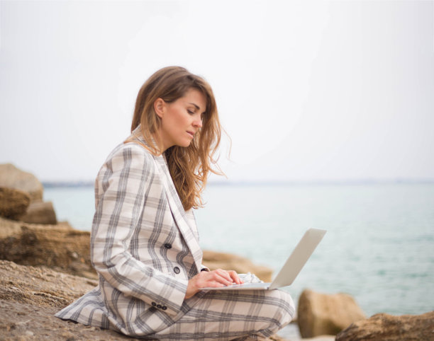 一位年轻女子坐在海边的一块石头上，手里拿着一台笔记本电脑。远程工作的概念图片下载