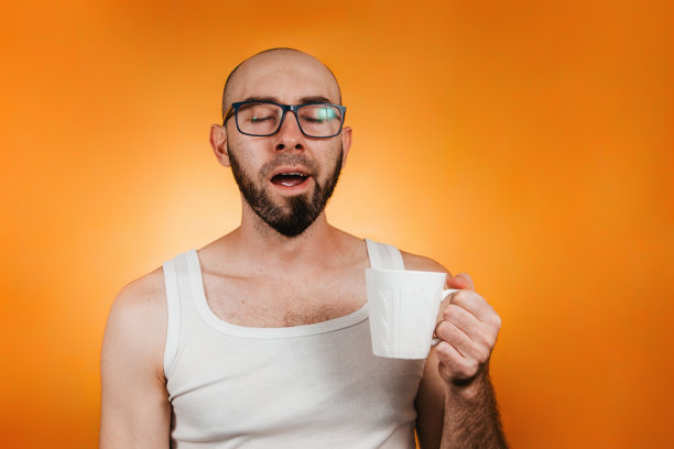 早起和工作效率的概念。一个秃顶，留着胡子，戴着眼镜，端着一杯咖啡或茶，懒洋洋地打着哈欠的男人。橙色背景。本空间图片下载