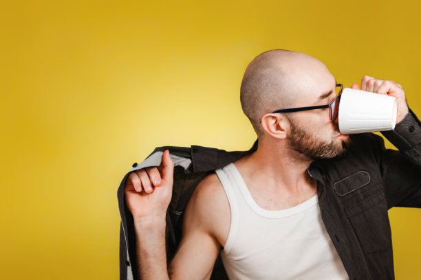 早上醒来和工作效率的概念。一个秃顶，留着胡子，戴着眼镜，穿着衬衫，同时喝着咖啡或茶的人。黄色背景。本空间图片下载