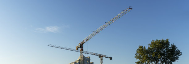 在城市中心的建筑工地上，一对高大的黄色起重机用来吊起沉重的建筑材料和蓝天背景下的树木。建筑行业。空间的文本。图片下载