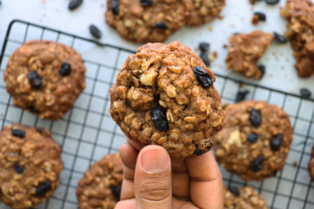 照片中，一个不认识的人拿着一块饼干，上面是一排排新鲜烘焙的自制燕麦和葡萄干饼干，放在金属丝冷却架上，周围是面包屑和葡萄干，大理石效果的背景，高架的视野图片下载