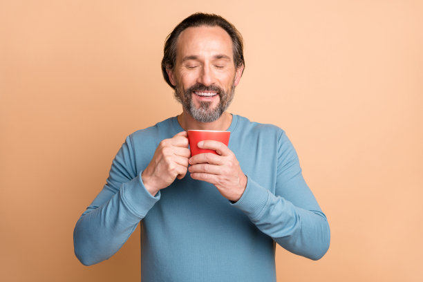 照片中的帅哥端着杯子闭着眼睛闻着味道穿着蓝色毛衣孤立的米色背景图片下载