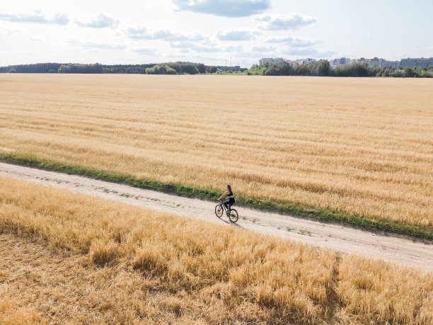 在一个阳光明媚的夏日，一名年轻女子骑着自行车沿着路边骑着小黑麦，背景中可以看到城市的房屋。后面的照片。图片下载
