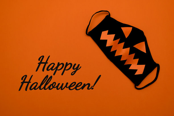 黑色保护面具，橙色背景上有橙色的眼睛和嘴巴。橙色背景上写着快乐的万圣节字样。万圣节和covid-19概念。图片下载