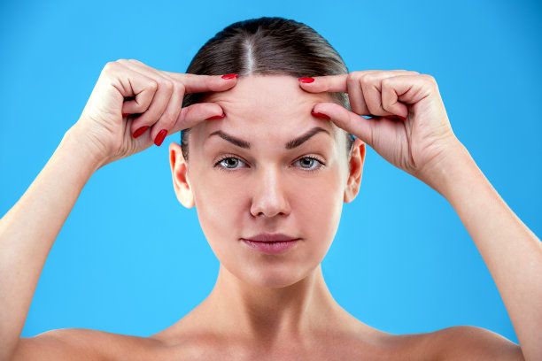 皮肤老化的迹象。一名震惊的年轻女子在蓝色背景下抚摸额头上的皱纹，失望地看着镜头图片下载