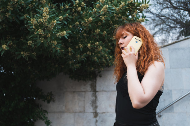 一个红发白种人女人站在水泥背景和绿树下打电话图片下载