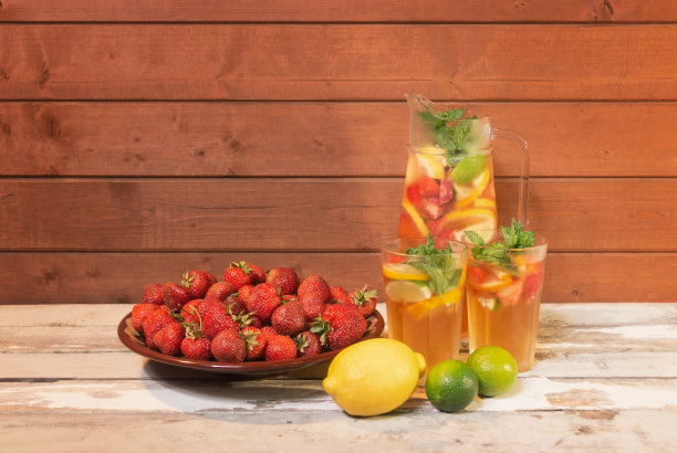 清凉的夏季柠檬汁，柠檬、草莓和酸橙装在水罐和玻璃杯里，放在陈年木桌上。附近有柠檬，酸橙和一盘草莓。图片下载