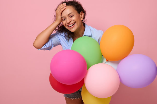 快乐的年轻混血美女笑着，玩着五颜六色的气球，孤立在粉红色的背景和复制空间。生日派对，庆祝活动的概念图片下载