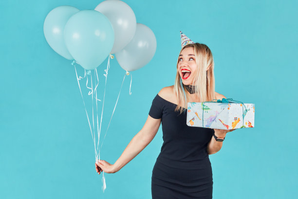 美丽微笑的女人身穿黑色连衣裙，头戴生日帽，手里拿着礼品盒和蓝色背景下的彩色气球。可爱快乐的年轻女孩庆祝生日派对。图片下载