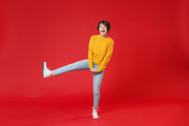 全长兴奋愉快有趣的年轻黑发女人20多岁穿着基本休闲黄色毛衣站起腿保持嘴张开孤立在明亮的红色背景摄影棚肖像。图片下载