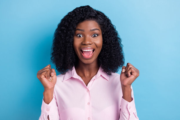 照片中的滑稽幸运深色皮肤的女人穿着粉红色的衣服，张开嘴举起拳头孤立的蓝色背景图片下载