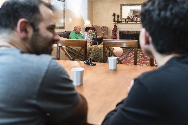 父亲和儿子围坐在餐桌旁，其他家庭成员，祖父母和一个十几岁的孙女，在背景中看电视。聚焦背景中的人，散焦前景。图片下载