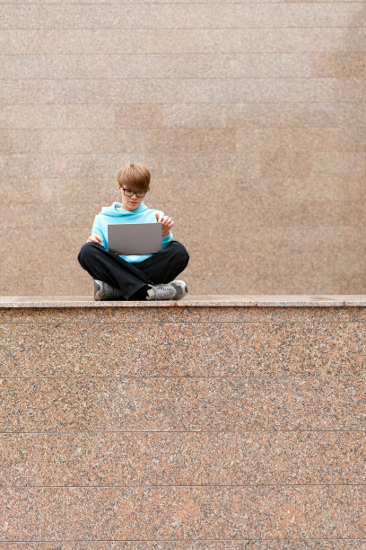 在城市环境中，一个留着短发的时髦女人拿着笔记本电脑的垂直照片。她坐在花岗石墙边的矮墙上，专心地工作着图片下载