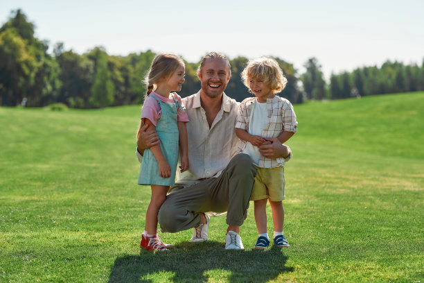 在夏季公园的草地上，快乐的爸爸和他可爱的小女儿一起站在草地上，对着镜头微笑图片下载