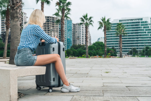 年轻苗条性感的金发女人穿着蓝色衬衫和灰色牛仔短裤坐在手机的长椅上，她的手靠在一个灰色的手提箱站在她的双腿之间，从后面的照片图片下载