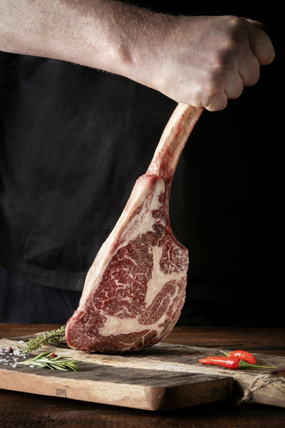 深色背景下，一名男子拿着一块生的战斧牛排和一根干的陈年牛肉骨头图片下载