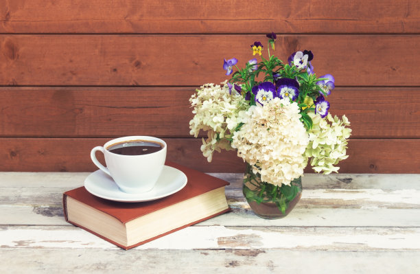一杯咖啡，一本书和白色绣球花和三色堇花在玻璃花瓶在旧木桌。图片下载