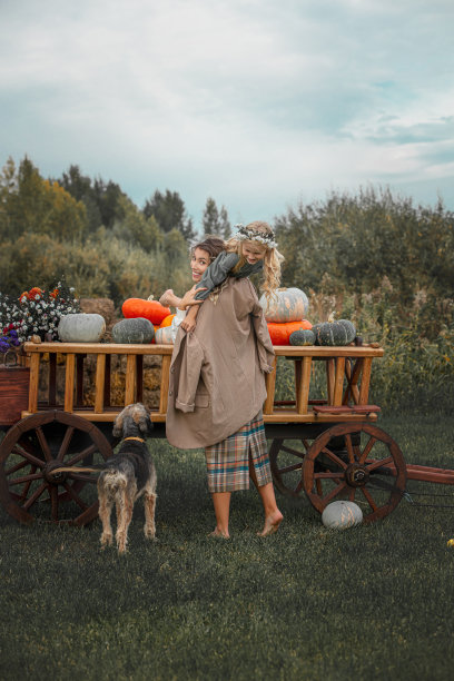 美丽幸福的家庭，妈妈、女儿和宠物狗在一起，坐在一辆装满五颜六色南瓜的木车上图片下载