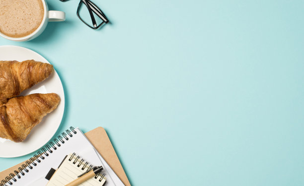 顶视图照片的工作场所的玻璃杯，杯子泡沫咖啡，笔提醒和盘子与两个羊角面包孤立的淡蓝色背景与空白图片下载