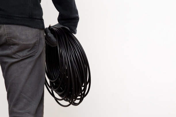 一个穿着深灰色衣服的年轻人。工人手戴纺织品保护手套，手持黑色卷电缆。在白色墙壁背景上为文本空出位置。特写镜头。后视图。图片下载