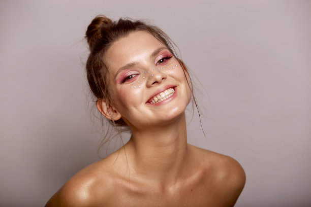 时尚创意化妆。年轻女子的面部表情与干净的新鲜的皮肤和明亮的妆与莱茵石雀斑在她的脸摆姿势在灰色的背景图片下载