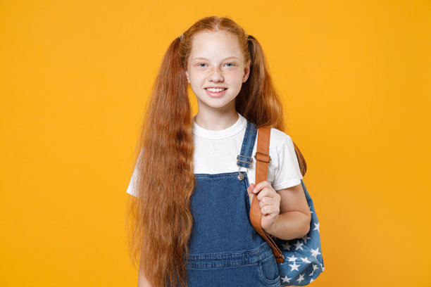 红发少女，12-13岁，扎着马尾辫，身穿白色T恤和蓝色牛仔校服，背着书包，在黄色背景前拍摄的儿童工作室肖像，展现儿童时期的教育生活方式图片下载
