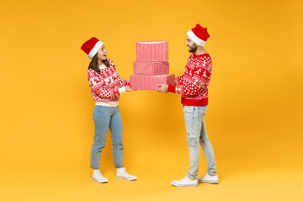 全长兴奋年轻的圣诞老人夫妇朋友男人女人在红色毛衣圣诞帽持有礼物盒与礼物孤立在黄色背景。新年庆祝快乐节日概念。图片下载