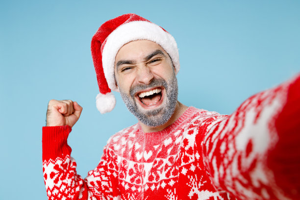 近距离拍摄快乐的北方大胡子男子冰冻的脸，戴着圣诞帽，穿着圣诞毛衣，用手机自拍，在蓝色背景上显示胜利者的姿态。冬时间概念新年快乐。图片下载
