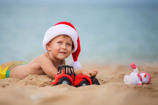 戴着圣诞老人帽子的快乐男孩在海滩上玩玩具车。圣诞节的孩子在海边休息。本空间图片下载