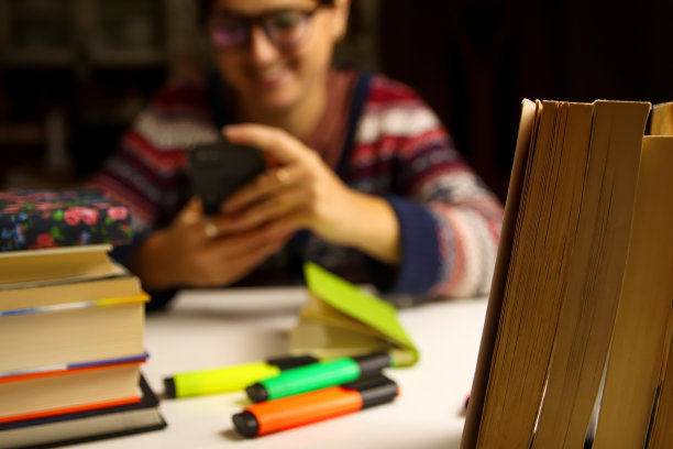 教育背景和学习观念模糊。年轻微笑的女人在晚上学习。千禧一代的学生在玩手机。准备考试。书和标记在前景上。前景聚焦图片下载