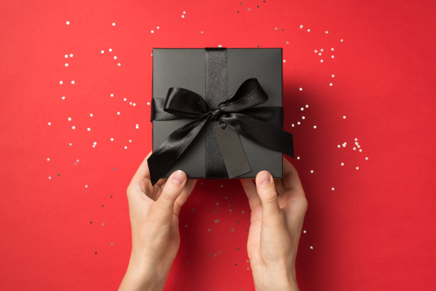 第一人称俯视图照片的女孩手握黑色礼盒与黑色丝带蝴蝶结和标签亮片孤立的红色背景与copyspace图片下载